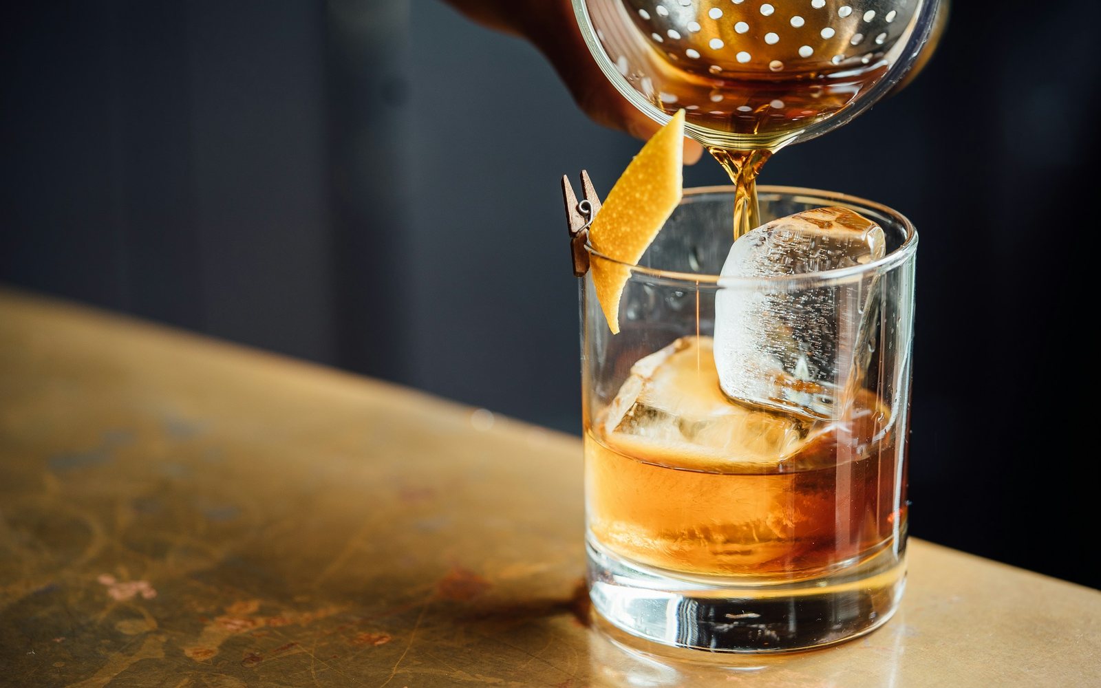 An Old Fashioned being poured over a large ice cube on a brass bar top, with an orange twist. Stock photograph, not this restaurant.